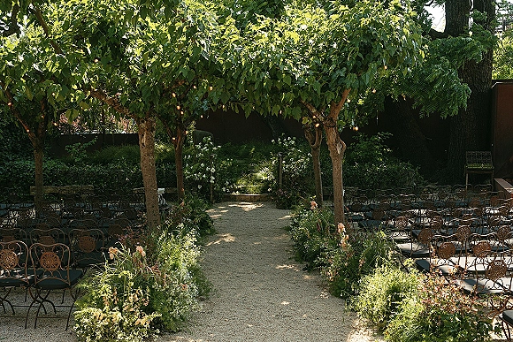 Ceremony aisle decor for an outdoor ceremony aisle with pastel flowers and greenery along a gravel path beneath a tree canopy and string lights