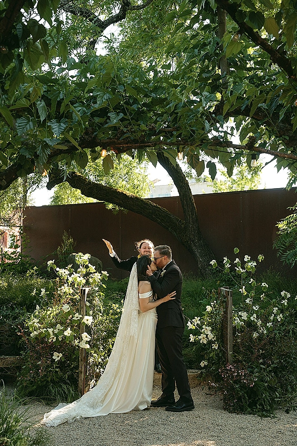 Wedding kiss portrait of bride and groom kissing, her long veil and train flowing under tree canopy with string lights in sunlit garden path