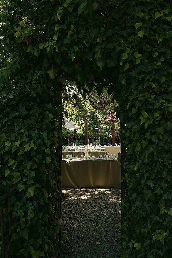 Outdoor reception setup with long banquet tables, neutral linens, glassware, and candlelight beside an ivy archway entrance in a garden pergola