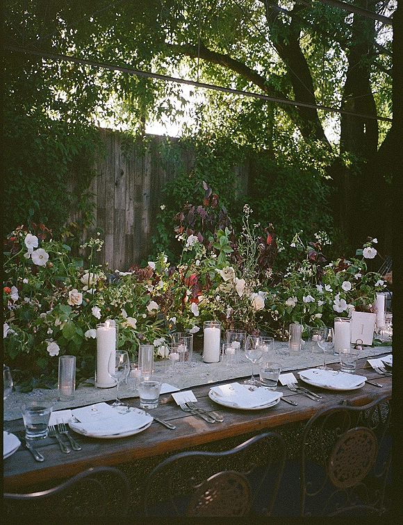 Reception tablescape with an outdoor reception table set with greenery garland, white flowers, candles, glassware, and string lights by an ivy fence