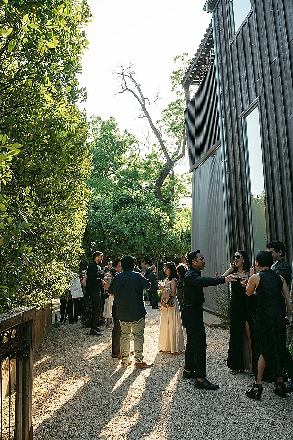 Wedding cocktail hour with wedding guests mingling in formal attire, holding drinks and wearing sunglasses in a sunny modern courtyard