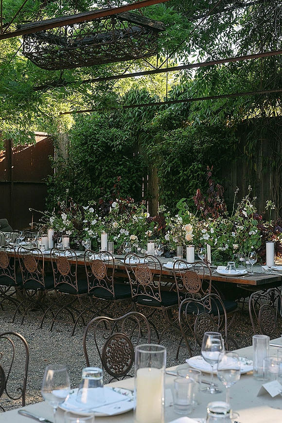 Reception tablescape with an outdoor reception table set with white taper candles, greenery garland, floral centerpieces, and wrought iron chairs in a garden pergola setting