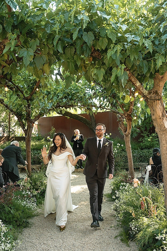 Wedding recessional as bride and groom walking hand in hand down a gravel aisle with string lights and white florals under a leafy canopy