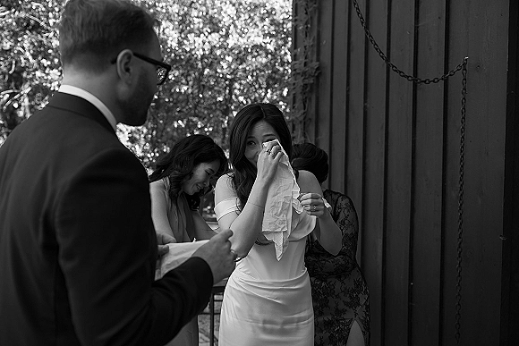 Bride emotional moment as she wipes tears with a handkerchief, wearing an off-the-shoulder gown while bridesmaids comfort her outdoors