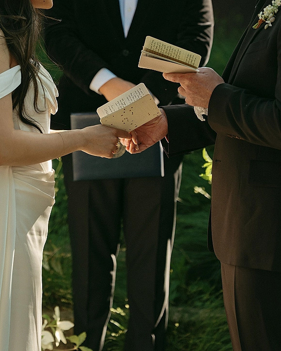 Wedding vows as the couple exchanging vows holds vow books and rings, bride in off-shoulder gown and groom in tux against greenery