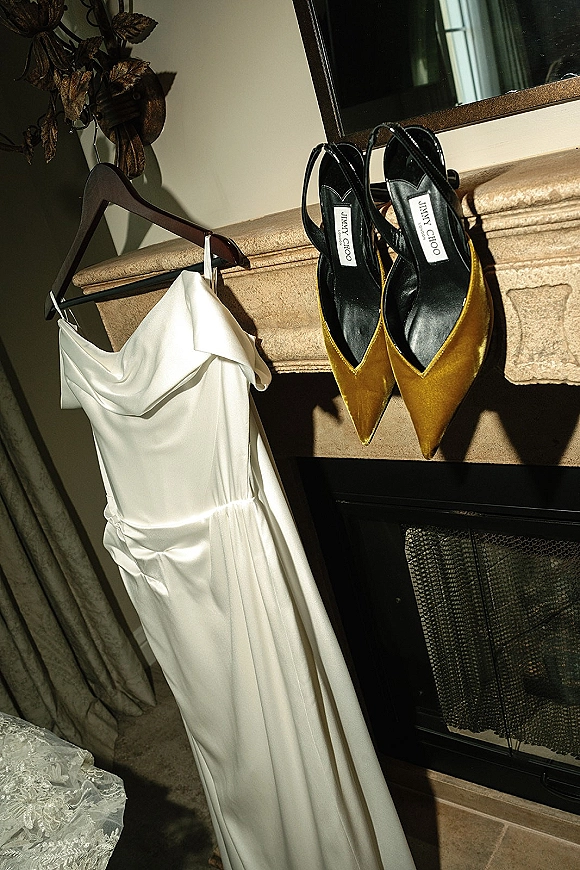 Wedding dress and shoes displayed by a fireplace mantel, bridal dress hanging on a hanger above pointed toe slingback heels, reflected in a mirror
