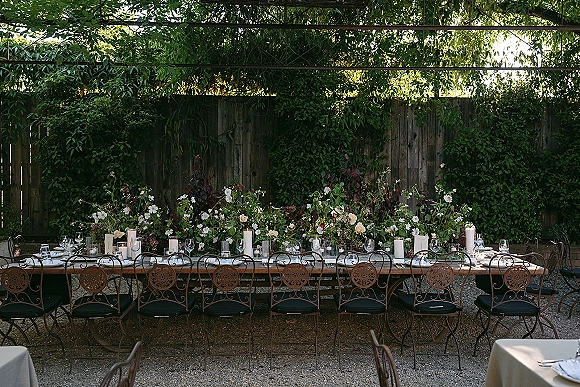Reception tablescape with a long banquet table dressed in white linens, floral garland, pillar candles, and place cards on a garden patio