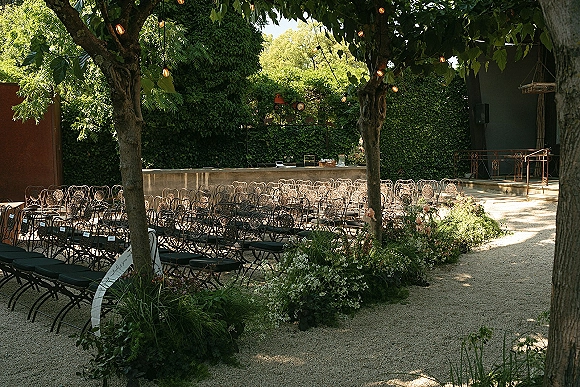 Outdoor ceremony setup with garden wedding ceremony seating in a semicircle, wrought iron chairs and aisle florals under string lights by an ivy wall