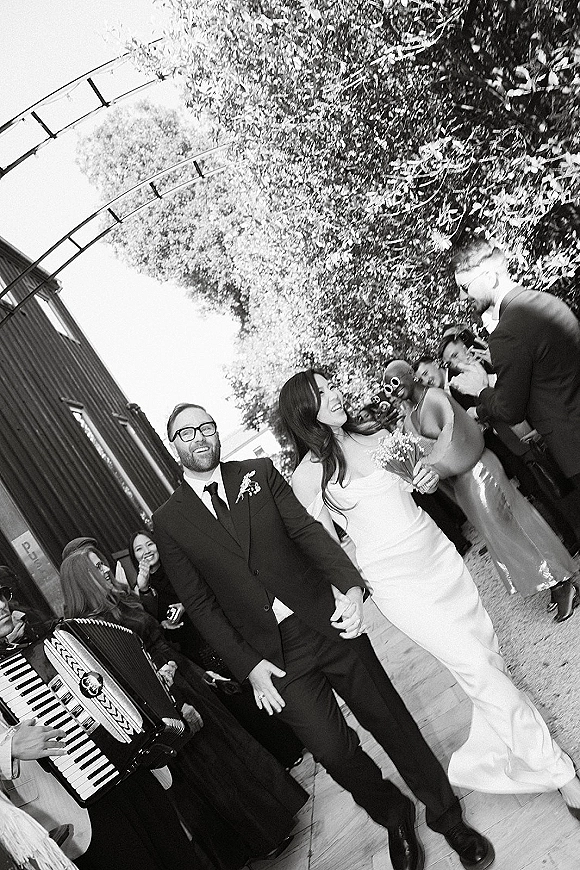Wedding recessional with bride and groom walking aisle hand in hand as guests cheer, confetti streamers fly on an outdoor walkway by trees