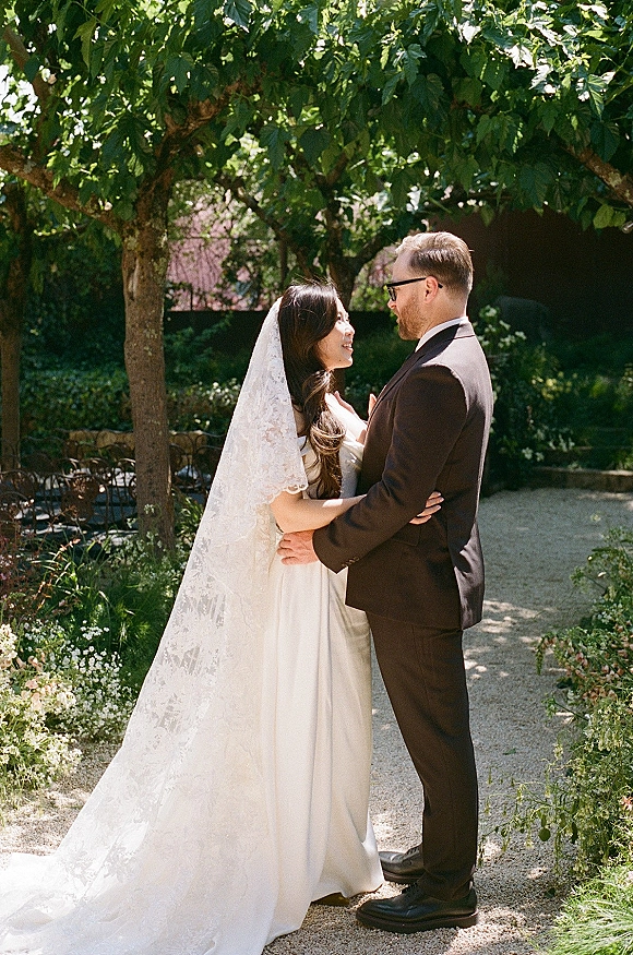 Couple portrait of bride and groom embrace on a gravel path under trees, bride in a wedding dress with lace veil, groom in suit and glasses