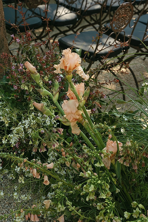 Wedding aisle florals with grounded aisle flowers in peach and white blooms with greenery lining a gravel path beside wrought iron chairs outdoors