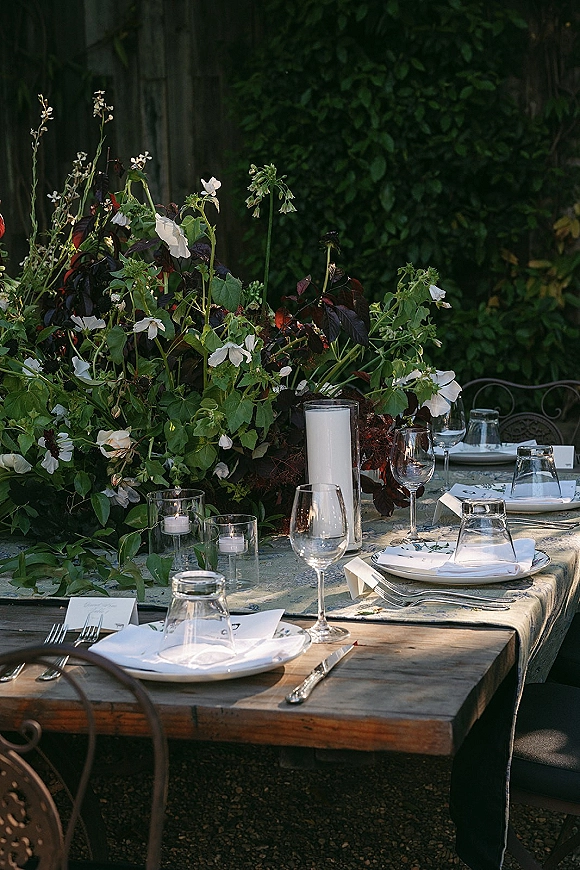 Reception tablescape with lush white floral and greenery centerpiece, candles in glass holders, and place settings on a patterned tablecloth outdoors