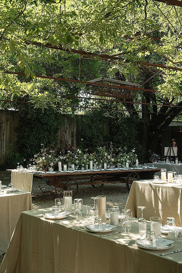 Reception tablescape with outdoor reception tables, white taper candles, glass cylinder candles, and greenery garland under a tree canopy