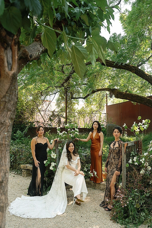Bridesmaid group photo of the bride with bridesmaids in mismatched dresses, bride seated in a lace veil on a garden path with greenery