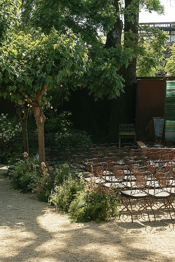 Ceremony setup for an outdoor wedding ceremony with wrought iron chairs lining a gravel aisle, floral ground arrangements, and string lights in trees near a fountain
