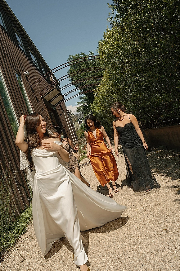 Bride with bridesmaids walking and laughing in satin wedding dress and veil, with mismatched dresses on a gravel path by a wood building