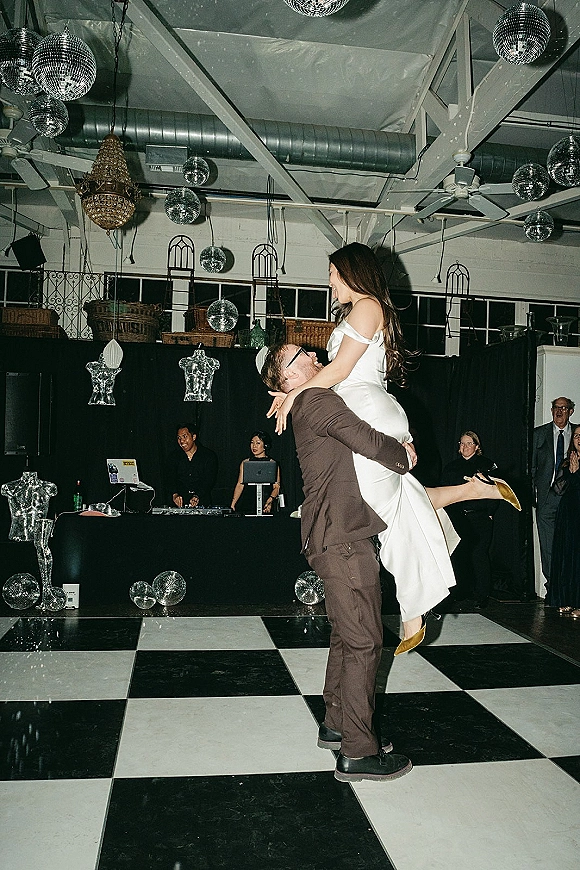 First dance moment as groom lifts bride in an off-shoulder dress on a checkered dance floor under disco balls and chandelier, guests watching