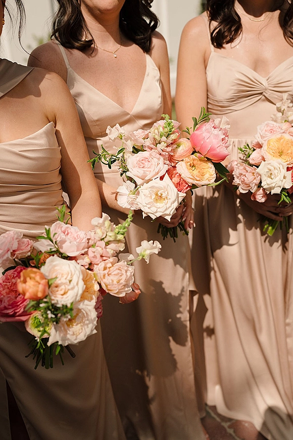 Bridesmaid bouquets with roses, peonies, and ranunculus held by women in blush bridesmaid dresses, sunlit outdoors with greenery accents