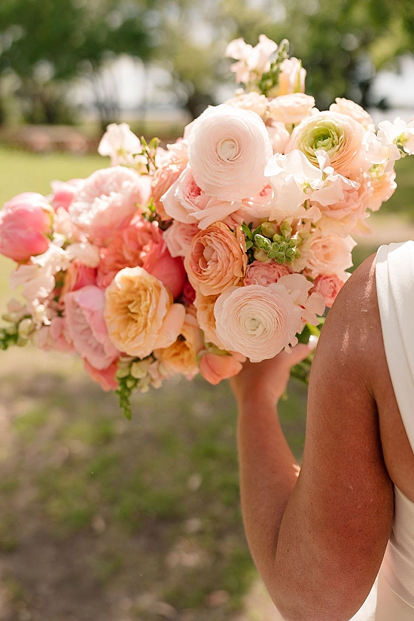 Bridal bouquet with ranunculus wedding bouquet blooms in peach and blush, held against a white bridal gown on a garden lawn with trees