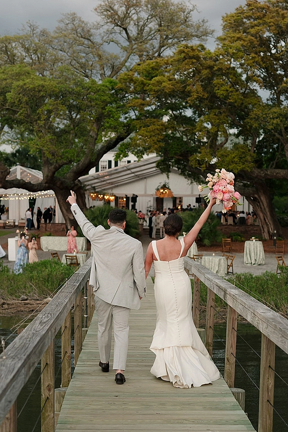 Wedding recessional as bride and groom walk away down a wooden dock, bride lifting a pastel bouquet overhead as guests cheer by the water