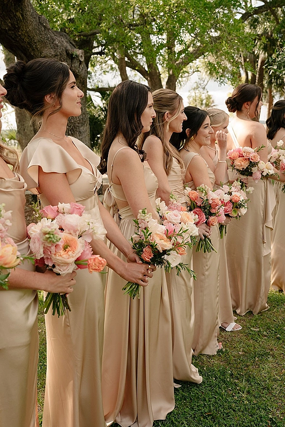 Bridesmaid lineup in champagne bridesmaid dresses holding pink and peach rose bouquets with greenery on a lawn under oak trees
