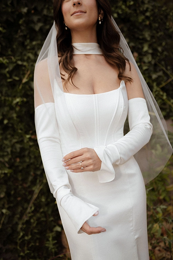 Bridal portrait of a bride in a strapless corset wedding dress with sheer veil, long gloves, and choker neckline against green garden foliage