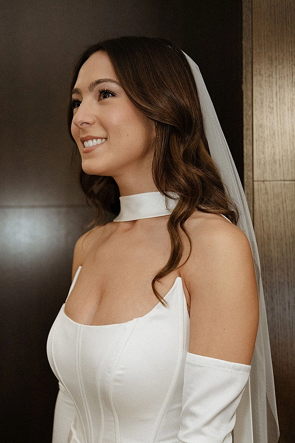 Bridal portrait of a smiling bride in side profile wearing a clean satin gown with tulle veil and gloves by an indoor doorway