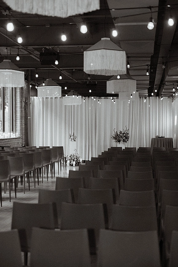 Ceremony setup with white draping and string lights over aisle chairs, floral clusters and candles in an industrial loft with brick walls