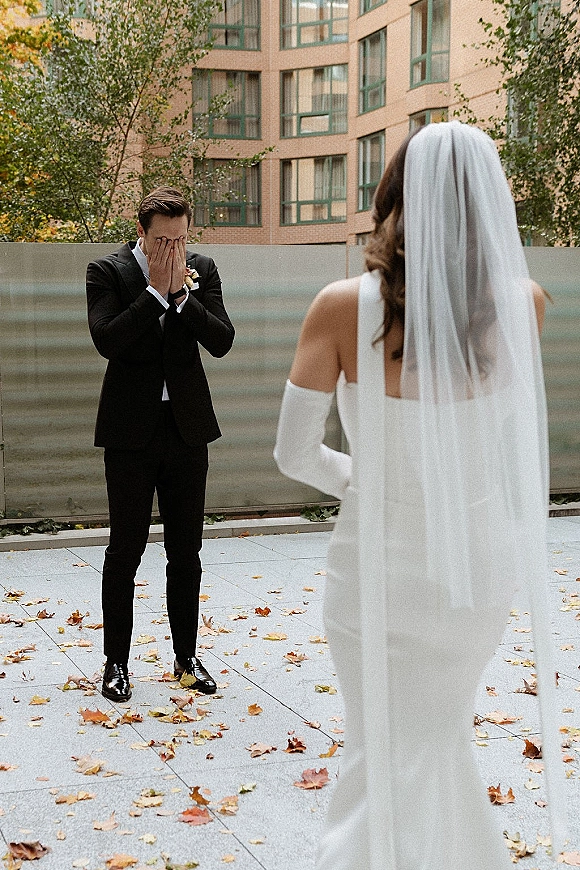Wedding first look as groom emotional reaction shows him covering his face while bride in long veil approaches in an outdoor courtyard