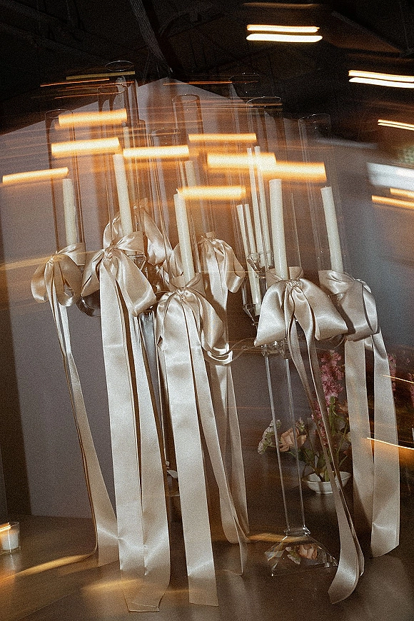 Wedding candle decor with a taper candle centerpiece in glass hurricane holders tied with ivory bows, glowing on a warm-lit reception table