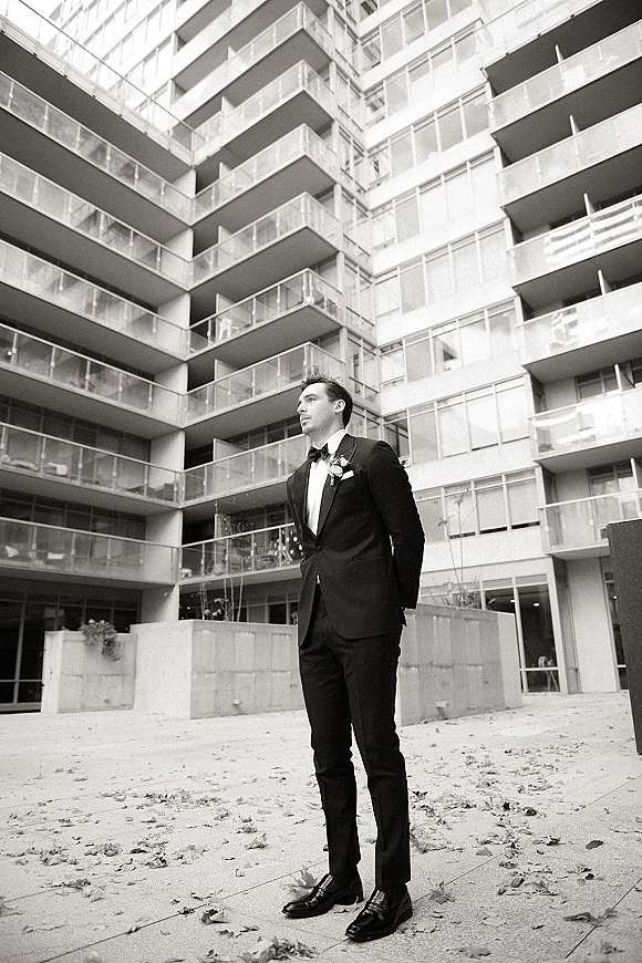 Groom portrait in tuxedo with bow tie and boutonniere, standing hands behind back in a modern apartment courtyard with leaves
