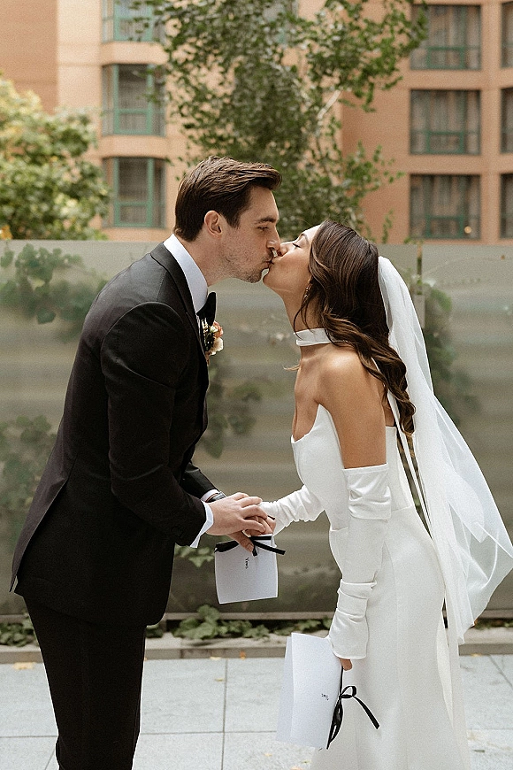 Wedding kiss portrait of bride and groom kissing, holding hands with vow books and ribbon on a city terrace with trees and glass railing