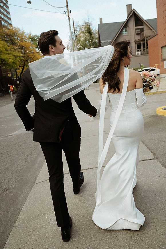 Couple portrait of bride and groom walking away holding hands, veil trailing as she carries a bouquet on a city street sidewalk