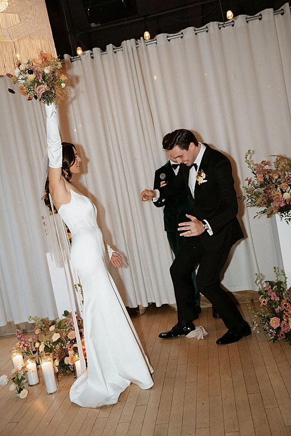 First dance at a wedding reception as bride in opera gloves raises her bouquet beside groom in tuxedo on a candlelit wood floor