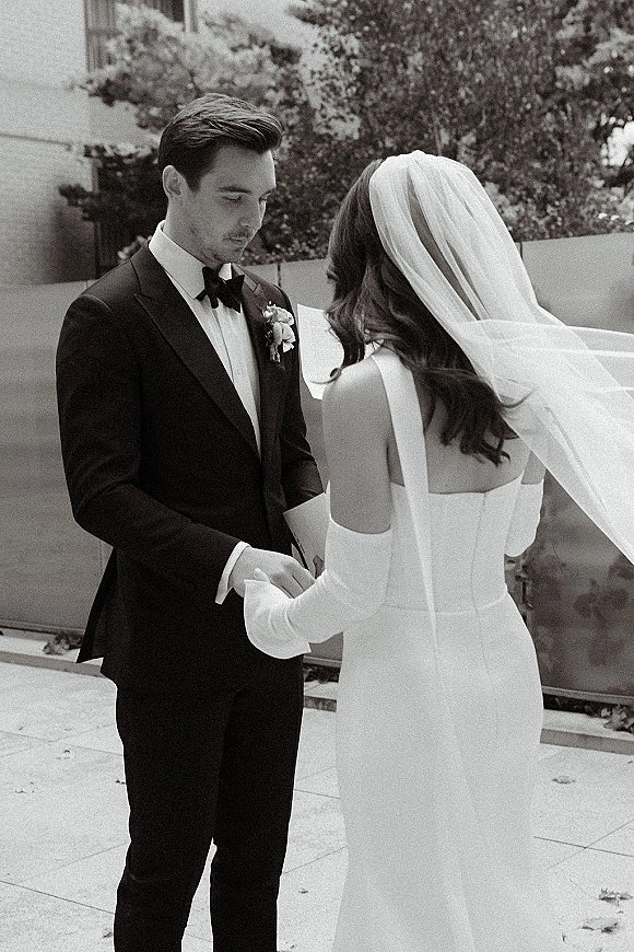 Wedding vows as bride and groom exchange vows from booklet, holding hands in an outdoor courtyard; veil and bow tie details visible