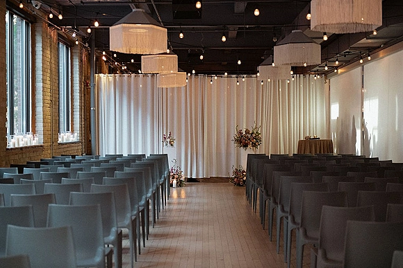 Ceremony setup for an indoor wedding ceremony with gray chairs lining an aisle runner, white drapery backdrop, and candles in an industrial loft