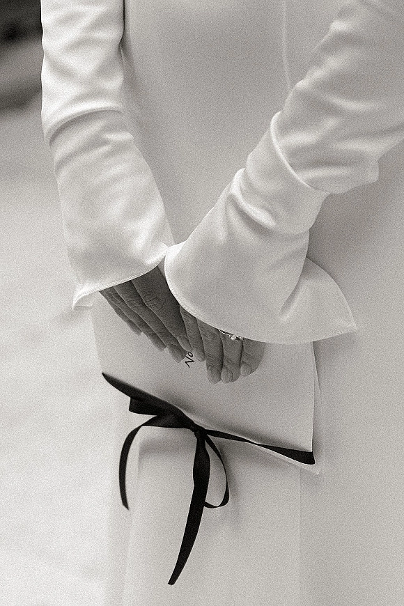 Wedding hands close-up with bride hands close up resting at waist, wedding ring visible beside wide white sleeves and black ribbon sash on light backdrop