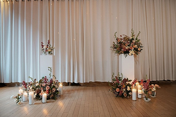 Wedding altar decor with pedestal floral arrangements and pillar candles on a wood floor, framed by white draping and curtain backdrop