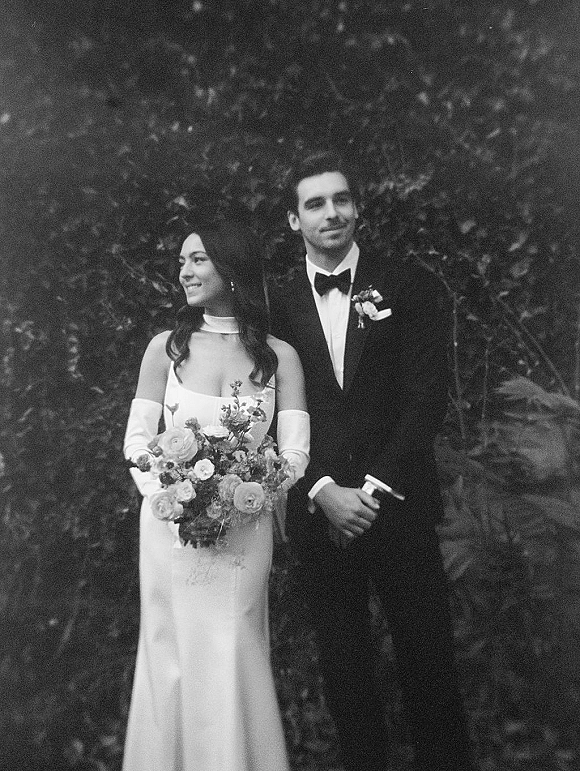 Couple portrait of bride in wedding dress and long gloves holding a bouquet beside groom in tuxedo with boutonniere, in garden foliage