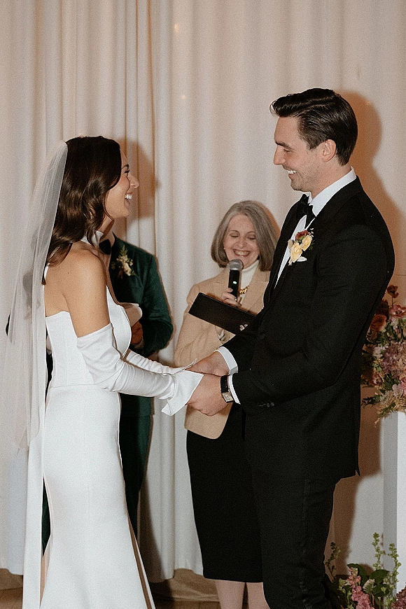 Wedding vows as bride in a strapless dress and veil holds hands with groom in a tuxedo before a white draped indoor backdrop