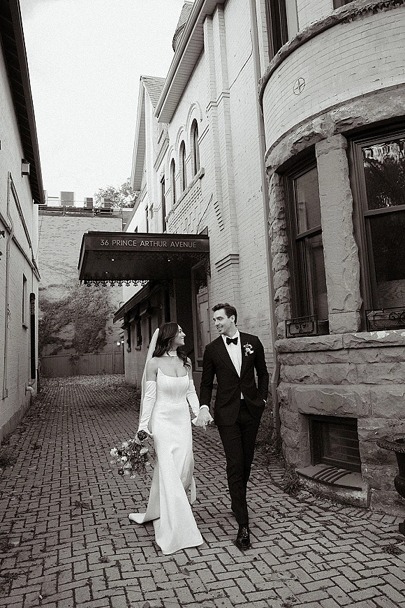 Couple portrait of bride and groom walking hand in hand, her veil and bouquet flowing, along a cobblestone street by brick buildings