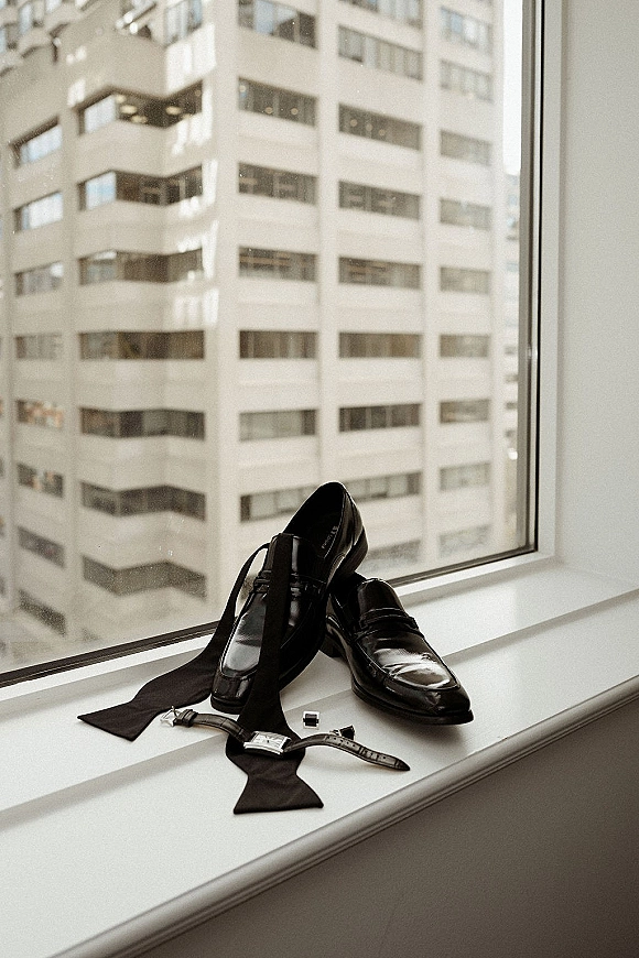 Groom accessories flat lay with black wedding shoes, bow tie, wristwatch and cufflinks arranged on a window ledge with city view