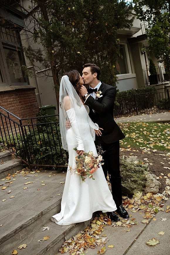 Wedding kiss portrait of bride and groom kissing on stone steps, bride in veil holding bouquet as fallen leaves line the sidewalk