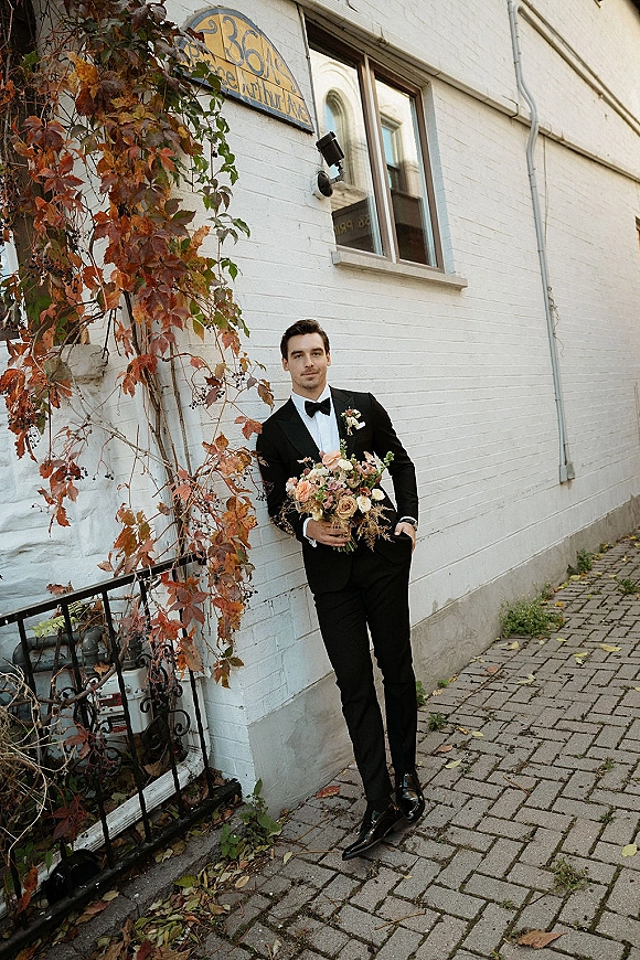 Groom portrait holding bouquet in a black tuxedo with bow tie and boutonniere, leaning by a white brick wall with ivy vines outdoors
