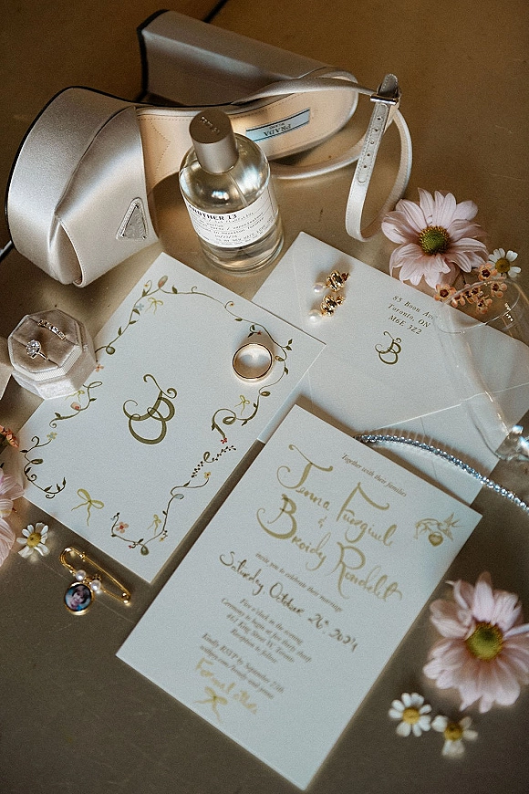 Invitation suite wedding invitation flat lay with calligraphy cards, wax seal envelopes, rings, pearl jewelry, heels, and daisies on a neutral tabletop