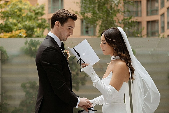 Wedding vows as the bride reads from a vow book with ribbon while the groom listens in an outdoor courtyard by concrete steps