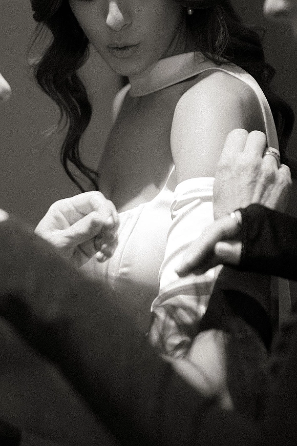 First dance moment as bride and groom sway close, her off-shoulder gown and wedding rings visible in moody reception light with blurred guests