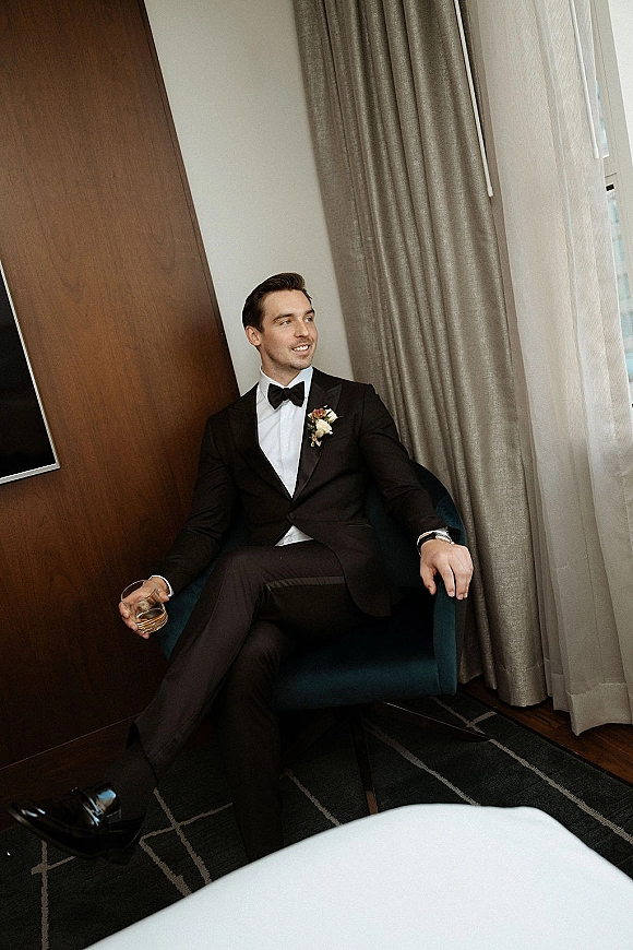 Groom portrait in a black tuxedo, seated in an armchair holding a whiskey glass, boutonniere and watch lit by a hotel window