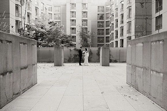 Couple portrait in a black and white wedding portrait style, bride in veil and tuxedoed groom holding hands in a modern courtyard