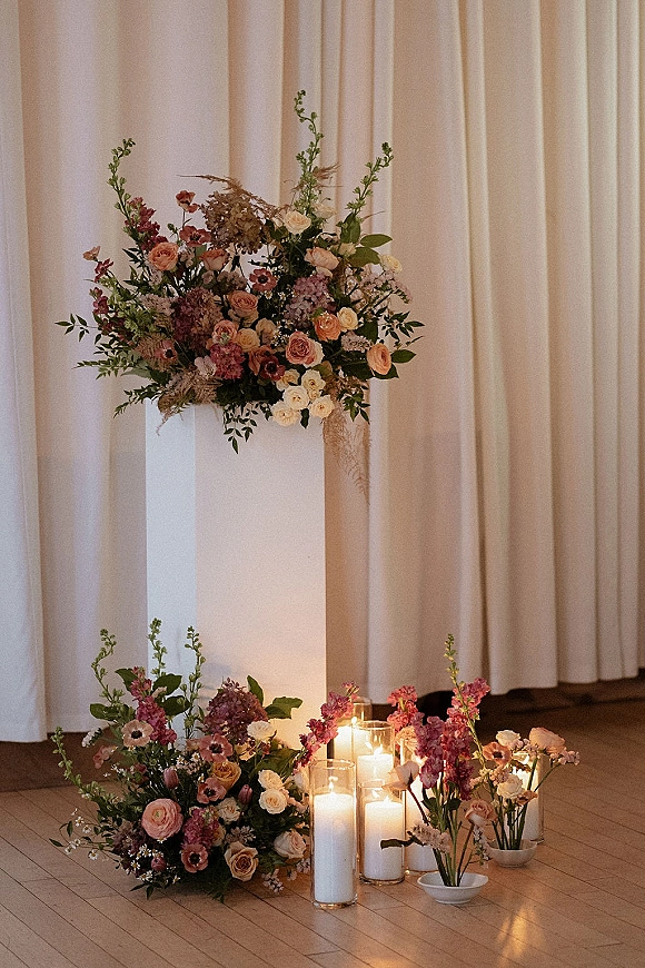 Wedding altar decor with ceremony altar flowers on a white pedestal, roses and anemones beside glass cylinder candles against white drapery